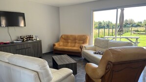 Un salon moderne avec un canapé blanc, un fauteuil en cuir marron et un canapé beige. Un téléviseur à écran plat est fixé au mur, il y a une table basse en bois et une vue sur un jardin à travers de grandes fenêtres.