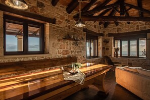 Interior rústico de piedra con un banco de madera, una mesa con un jarrón y vista al mar a través de amplias ventanas.