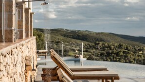 Un mur de pierre agrémenté d’un élément décoratif aquatique, de chaises longues et d’une piscine, le tout offrant une vue sur un paysage vallonné.
