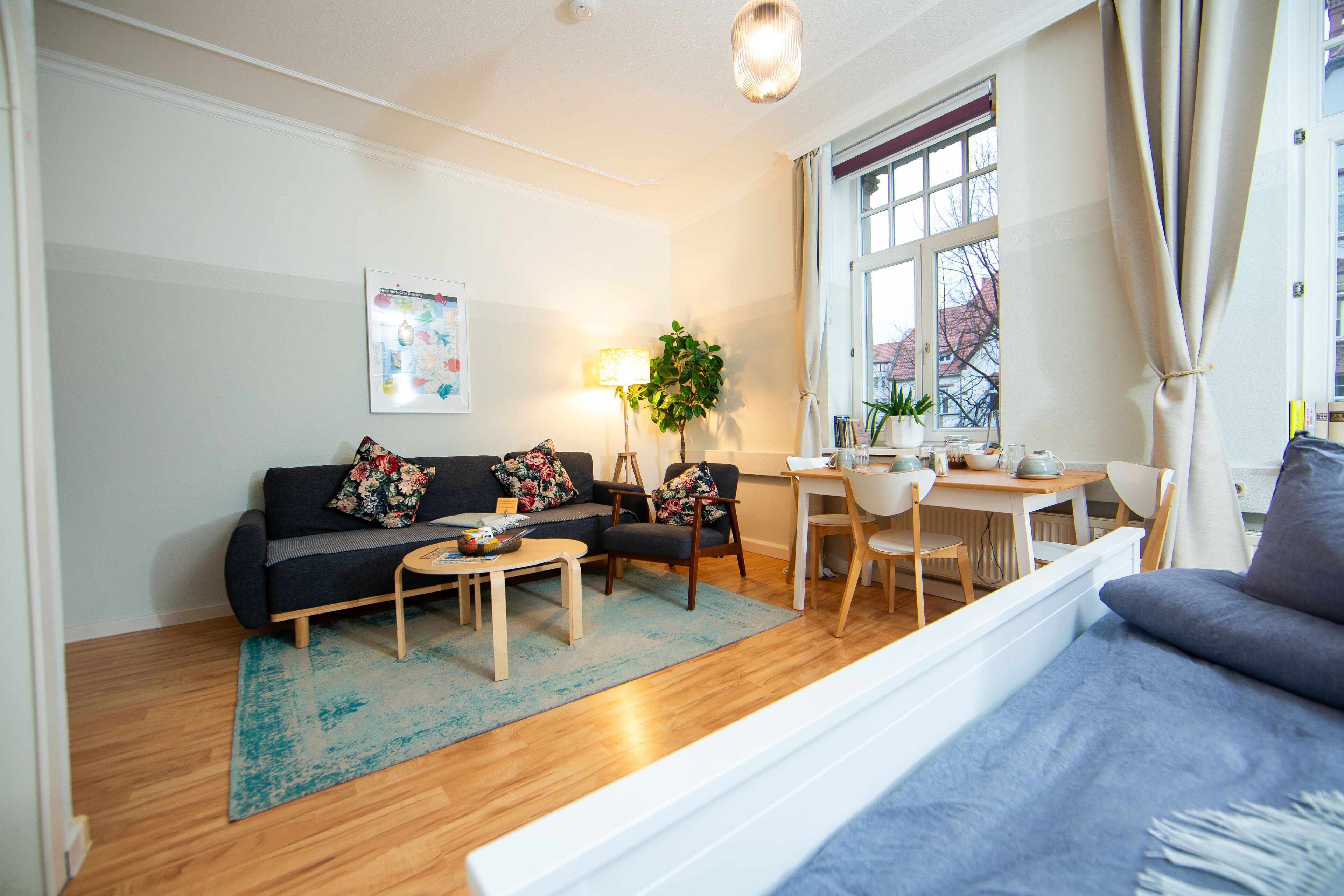 Ein Wohnzimmer mit einer Couch, zwei Sesseln, einem Couchtisch, einer Lampe sowie einem Essbereich mit Tisch und Stühlen.