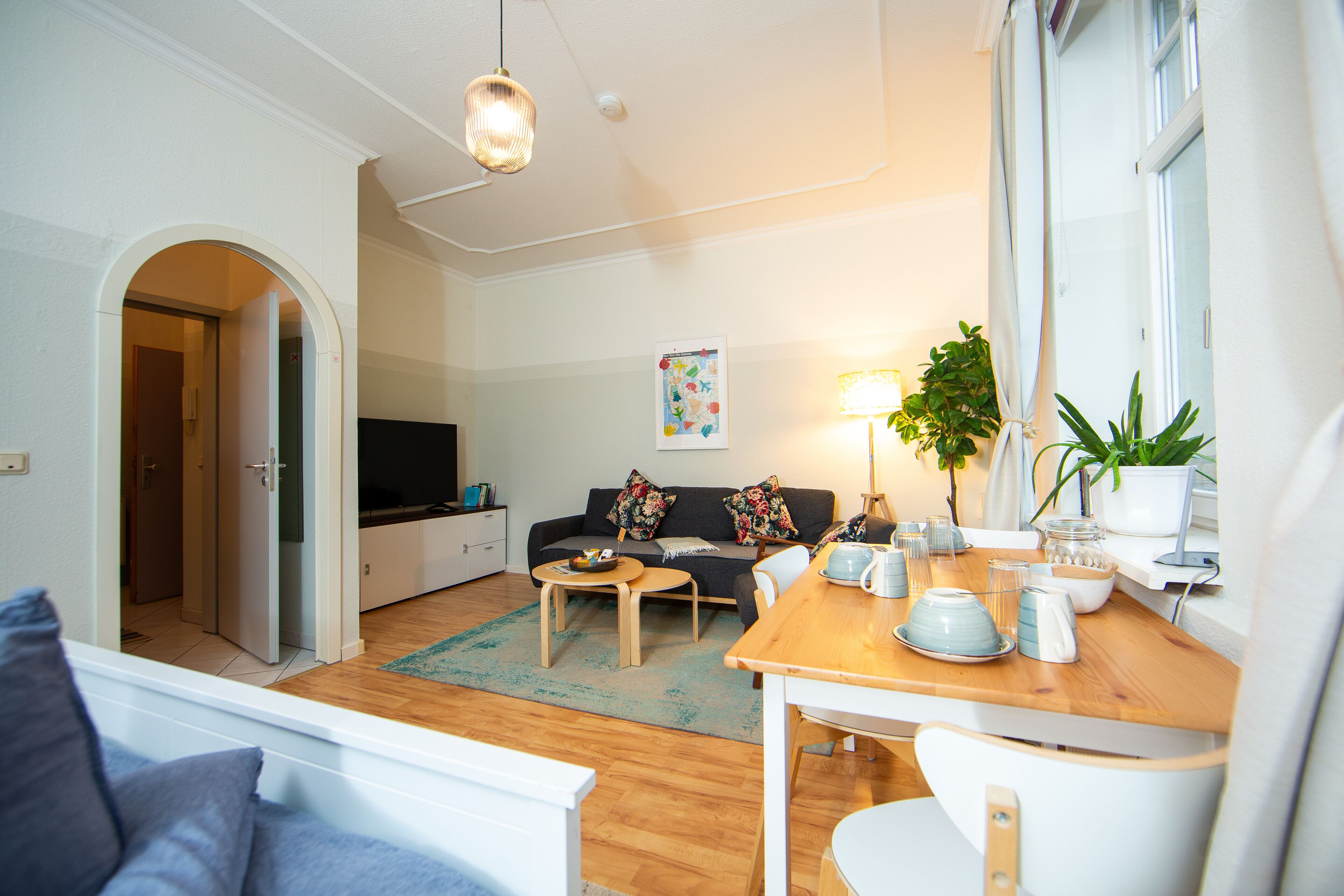 A modern living room with a sofa, coffee table, and dining table. There is a TV, a plant, and a window with curtains.