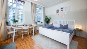 Un dormitorio con una cama blanca, un escritorio y sillas. Hay ventanas con cortinas, un radiador y una planta sobre la mesita de noche.