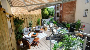 Un patio techado al aire libre con mobiliario de madera, plantas en macetas y vista a un edificio de ladrillo.