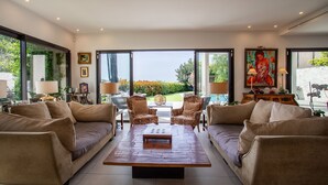A modern living room with two sofas, a coffee table, and two armchairs. There are large windows offering a view of a garden. - Villa Les Sources VI4245 By Riviera Holiday Homes (Niza)