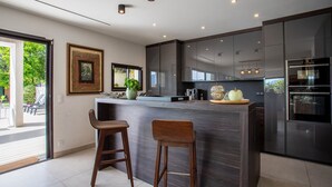 A modern kitchen with dark cabinetry, a central island with stools, and a large window providing a view of the outdoors. - Villa Les Sources VI4245 By Riviera Holiday Homes (Niza)