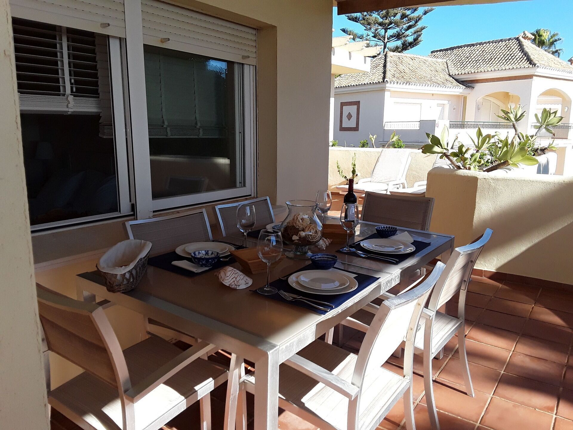 A dining table set for four with white chairs, a window with blinds, and a view of a residential area with a tiled roof and plants.
