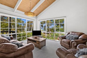 A modern living room with leather sofas, a coffee table, and a flat-screen TV. Large windows offer a view of greenery and trees.