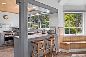 A kitchen with a central island, wooden stools, a built-in oven, and a window with a view of greenery.