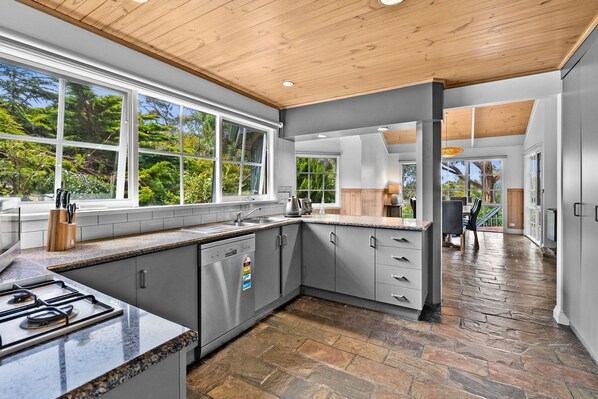 A modern kitchen with a stone tile floor, a built-in dishwasher, and a large window overlooking greenery.