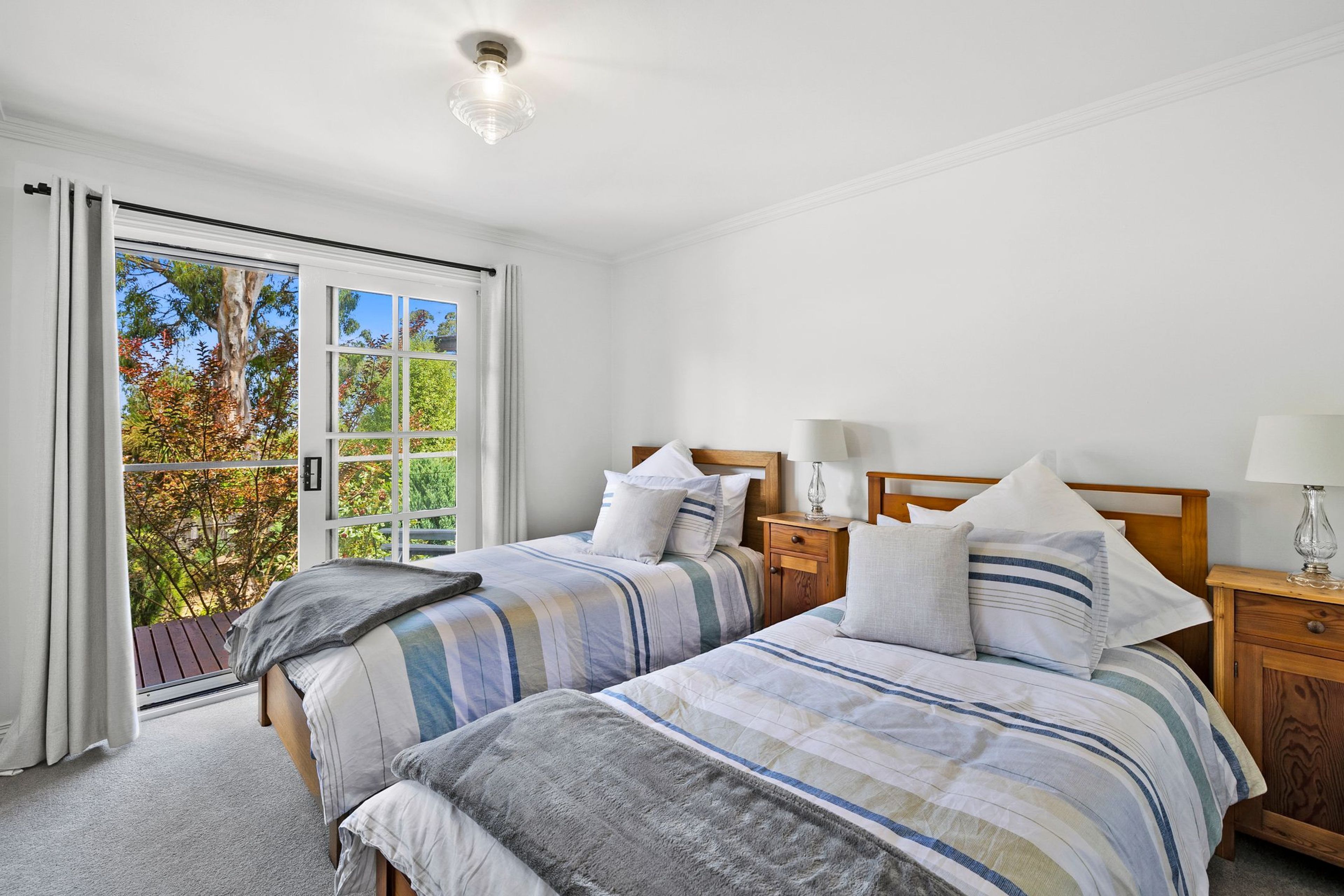 A bedroom with two beds, a window with curtains, a nightstand with a lamp, and a ceiling light.