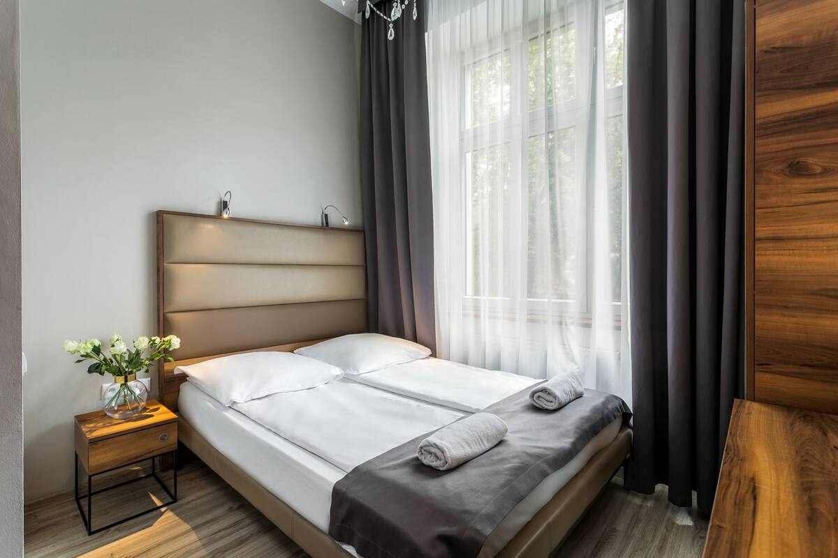 A neatly made bed with a wooden headboard, a bedside table with a vase of flowers, and sheer curtains on a window.