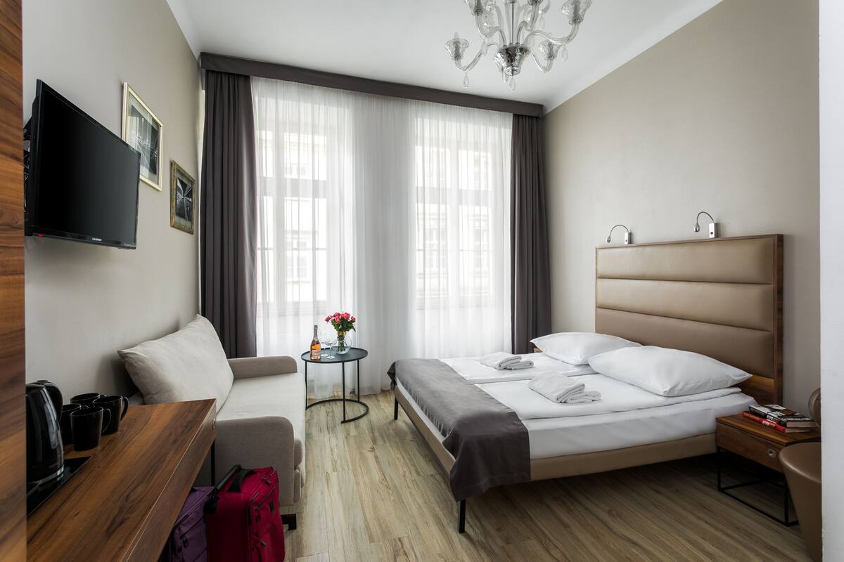 A modern hotel room with a bed, a sofa, a TV, a small table with a vase of flowers, and a wooden desk.