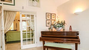 A piano with a bench in a room with a tiled floor, a door with curtains, and a wall with framed pictures and a wall sconce. - Swallows Barn (Chesterfield)
