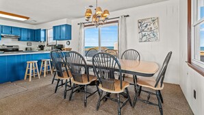 A modern kitchen with blue cabinets, a dining area with a wooden table and chairs, and a view of the ocean through large windows. - Dawn Patrol 4 Bedroom Home in Kitty Hawk (Kitty Hawk)