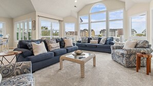 Un salón amplio con techo alto, ventanas grandes y vistas a edificios. Hay dos sofás y un sillón con cojines estampados.