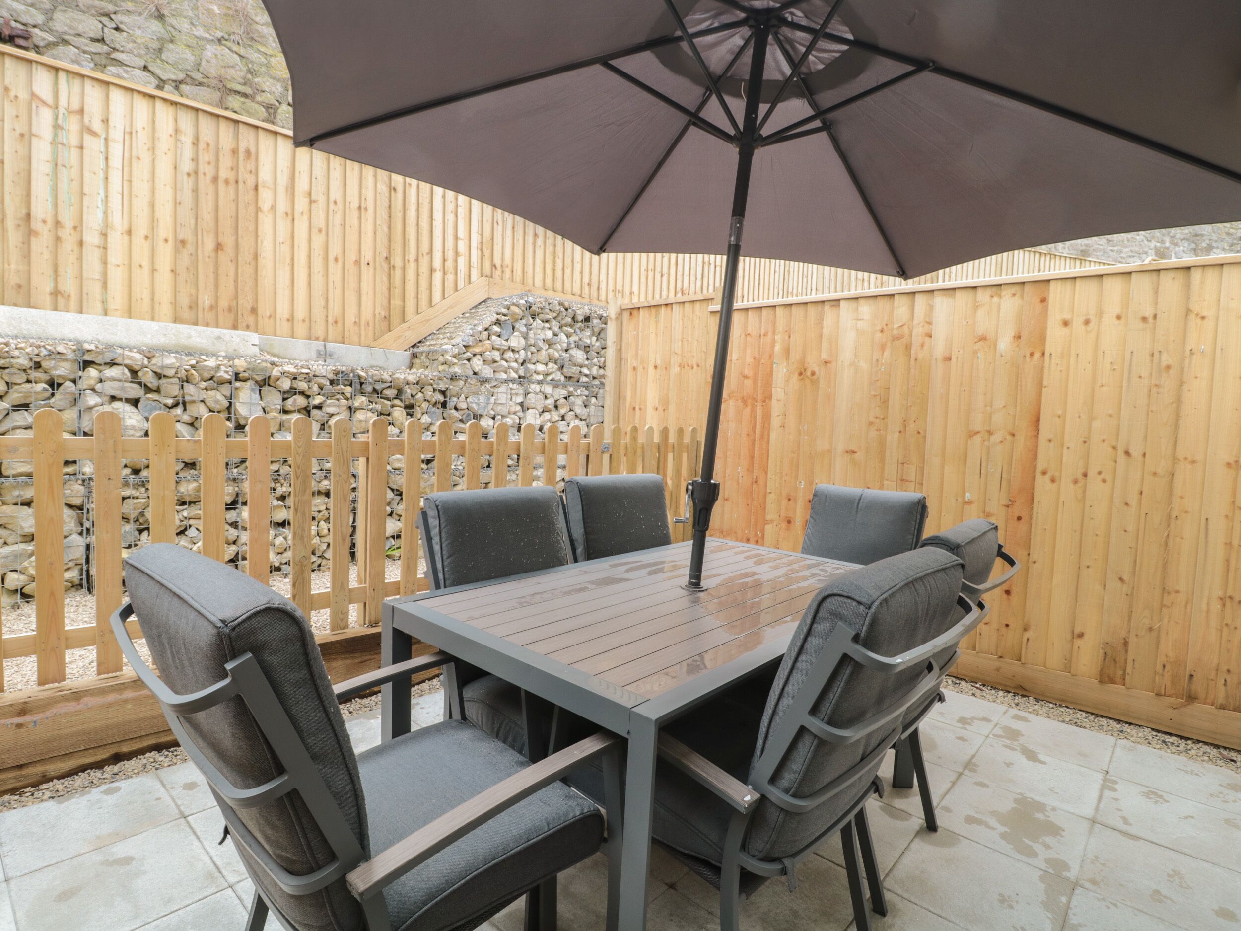 An outdoor patio with a wooden table and six chairs, an umbrella, and a stone wall in the background.