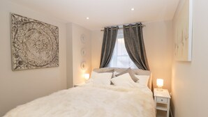 A bedroom with a large bed, a nightstand, a wall-mounted lamp, and a window with curtains. - Kimberlin (Portland)