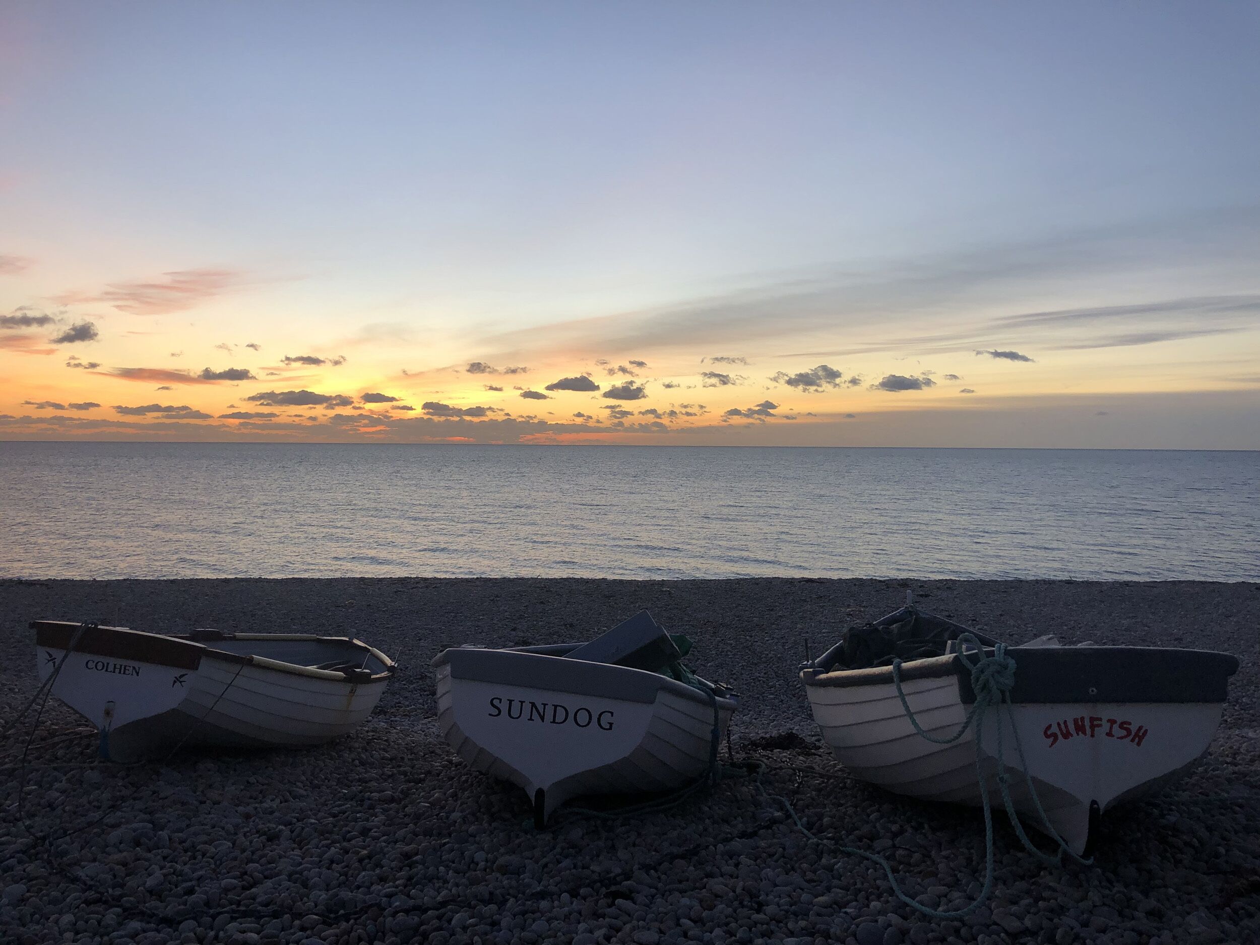 Τρία σκάφη, με ονόματα COLHEN, SUNDOG και SUNFISH, βρίσκονται σε μια παραλία με βότσαλα, με το ηλιοβασίλεμα να σχηματίζει ένα εντυπωσιακό φόντο.