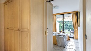 Quarto com um guarda-roupa de madeira grande, cama com manta cinzenta e vista para um jardim por uma porta de correr.