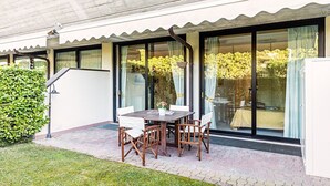Um pátio com mesa e cadeiras, um vaso de planta e vista para um jardim através de grandes portas de vidro deslizantes.