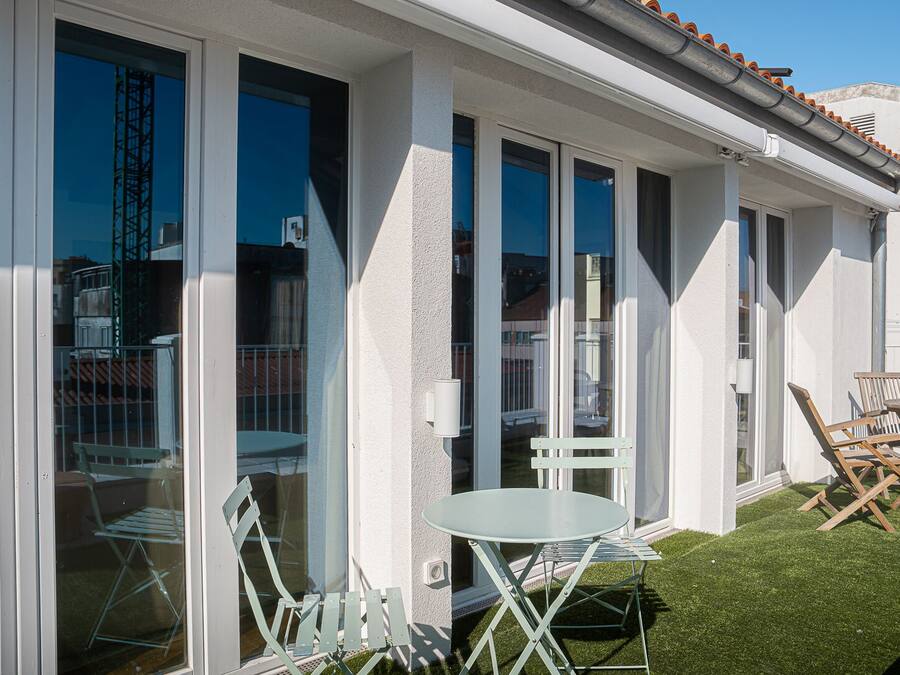Un patio techado al aire libre con mesa y sillas, un pequeño jardín y vistas de los edificios circundantes.