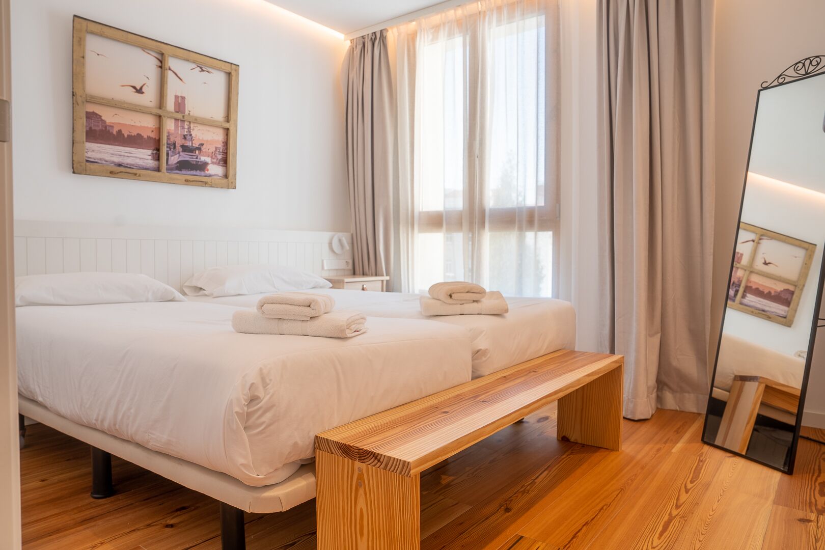 Une chambre moderne avec deux lits, un banc en bois, un grand miroir et un tableau représentant un paysage urbain accroché au mur.