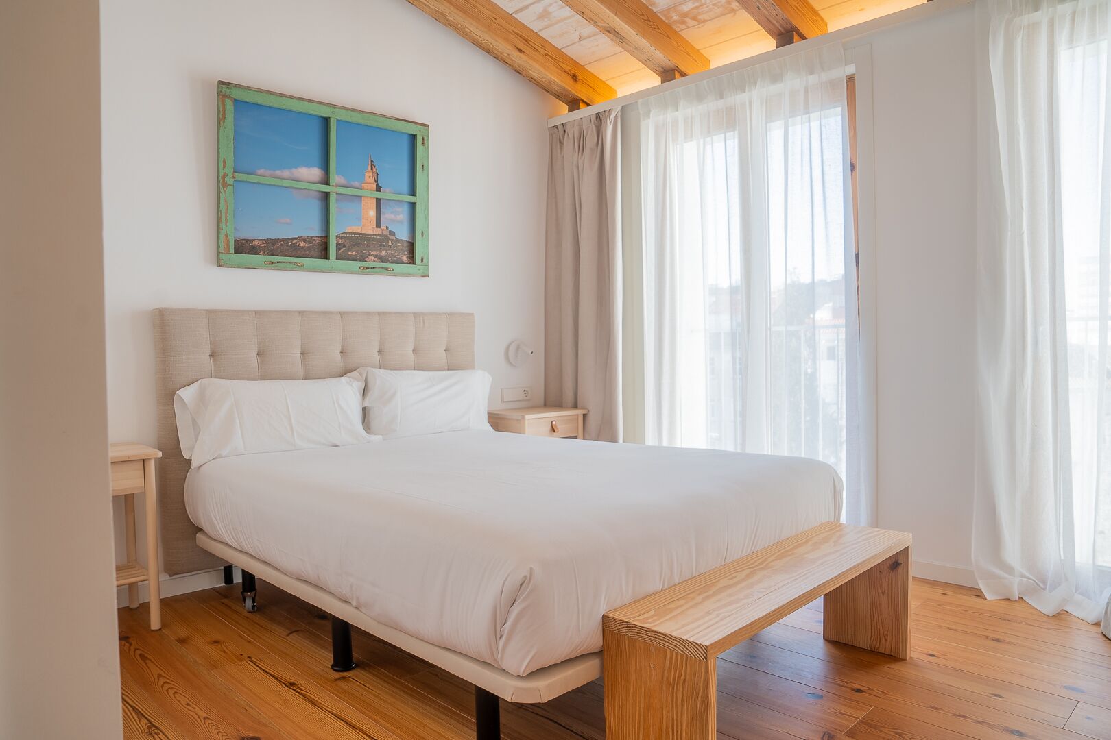 Un dormitorio ordenado con una cama grande, un banco de madera, una mesita de noche y una ventana con cortinas translúcidas.