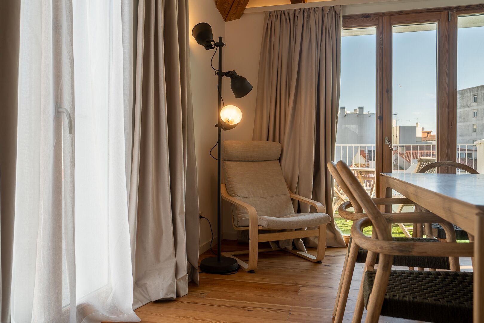 A room with a wooden floor, a beige armchair, a dining table with chairs, a floor lamp, and large windows with curtains.