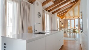 Een moderne keuken met een wit kookeiland, houten balken en uitzicht op een eethoek.