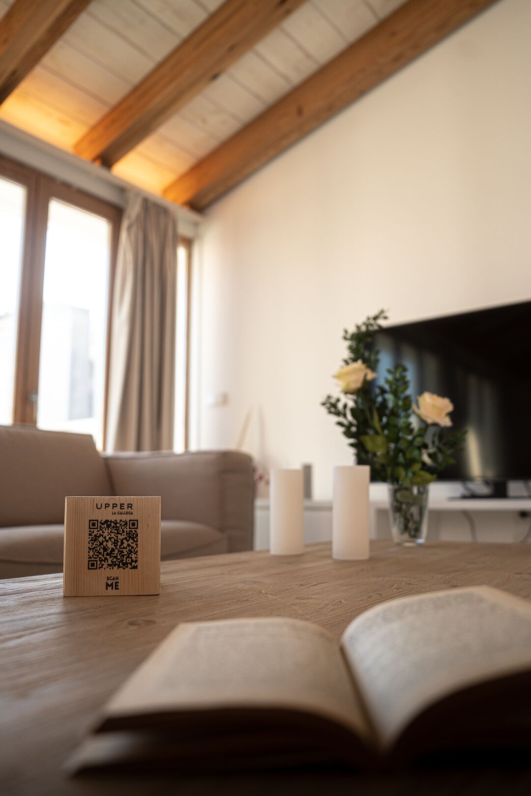 A cozy living room with a wooden ceiling, a sofa, a TV, a vase with flowers, and a QR code on a wooden box.