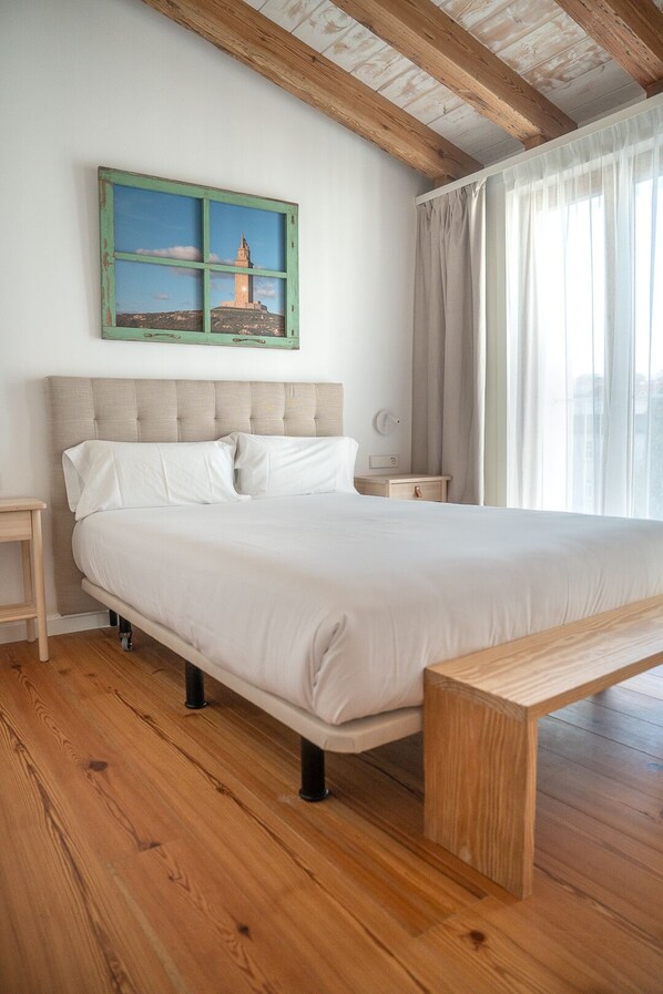 A bedroom with a large bed, wooden floor, a nightstand, a bedside table, a bench, a window with curtains, and a painting on the wall.