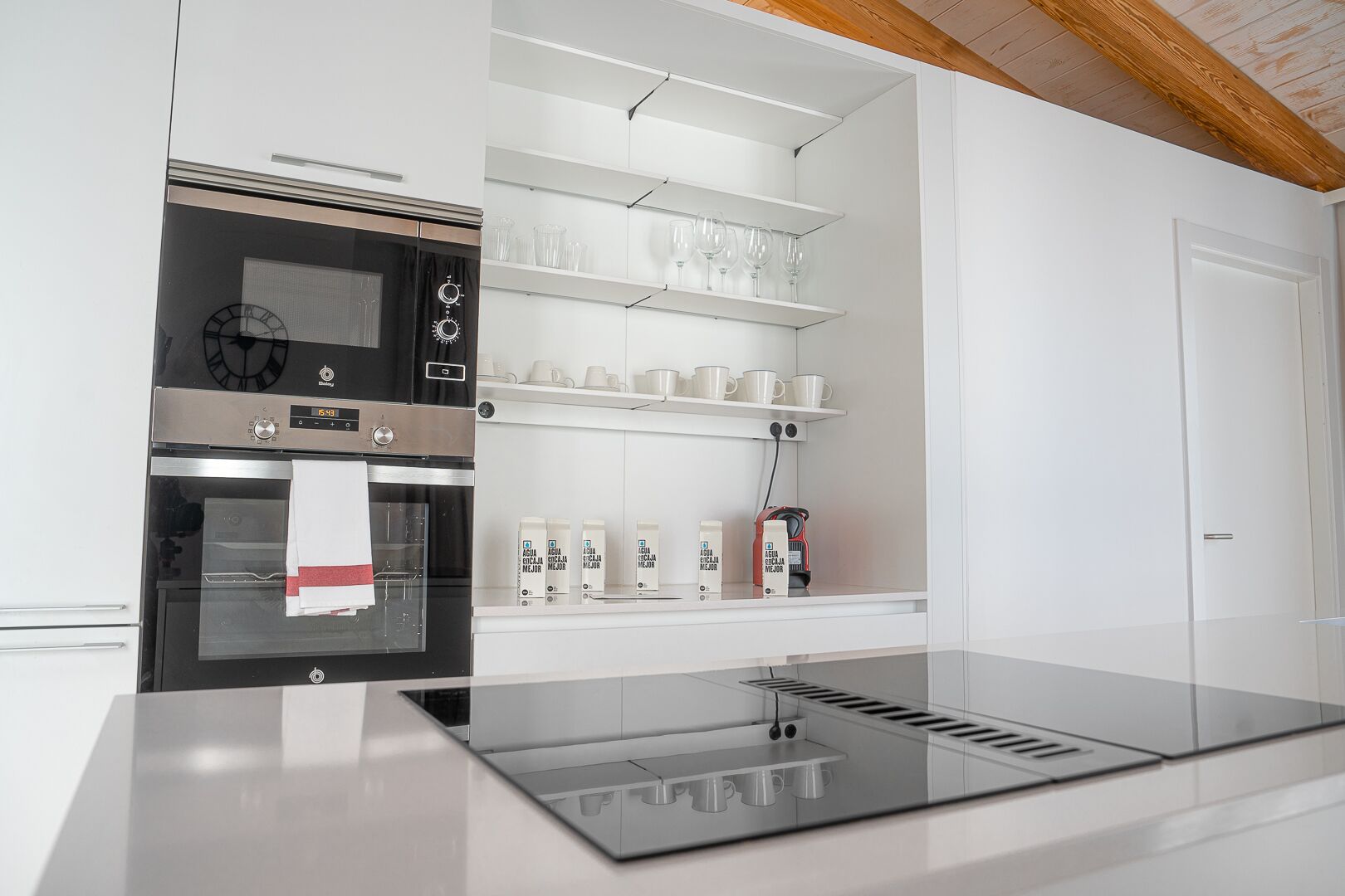 A modern kitchen with a built-in oven, a coffee machine, and shelves with cups and containers.
