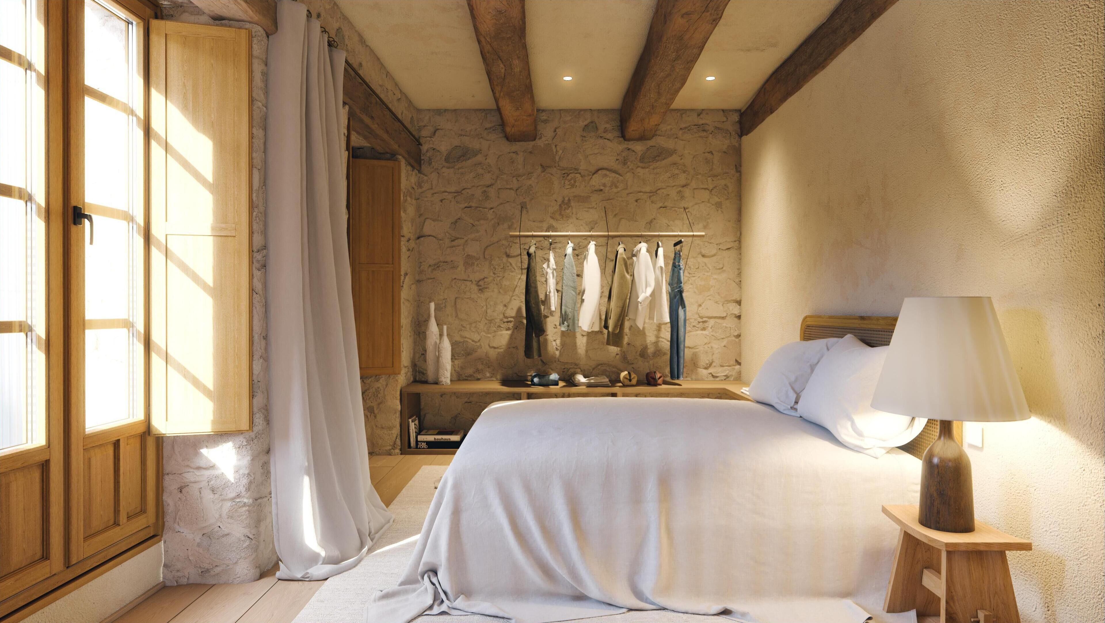 A bedroom with a stone wall, wooden beams, a bed with white linens, a nightstand with a lamp, and a clothing rack with various garments.