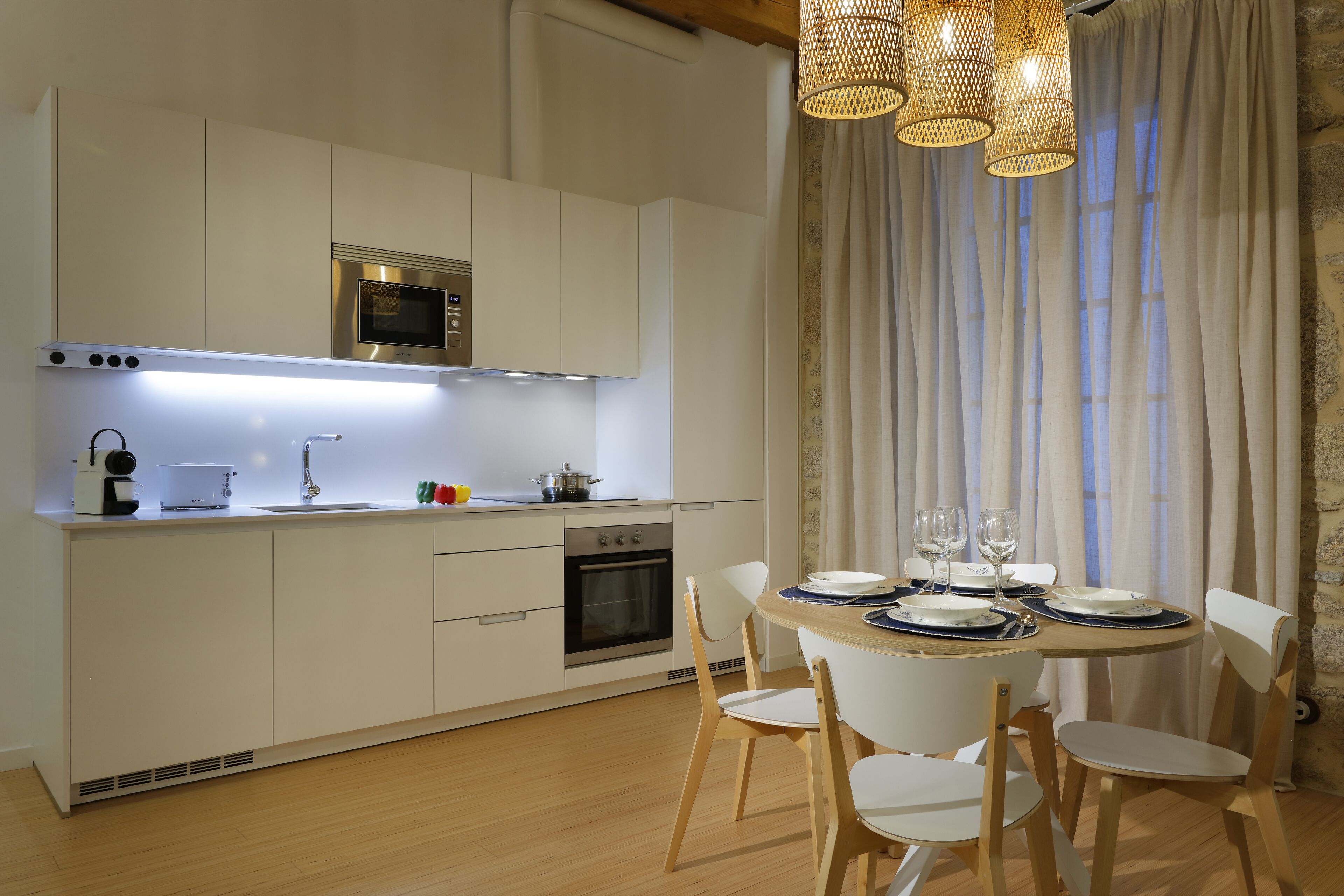 Cozinha moderna com armários brancos, forno embutido e área de refeições com mesa de madeira e cadeiras brancas.