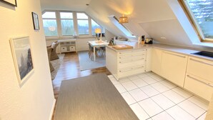 A modern kitchen with white cabinetry, a dining area with a table and chairs, and large windows allowing natural light. - Kieler FeWo No.1, kieler-fewo de (Kiel)