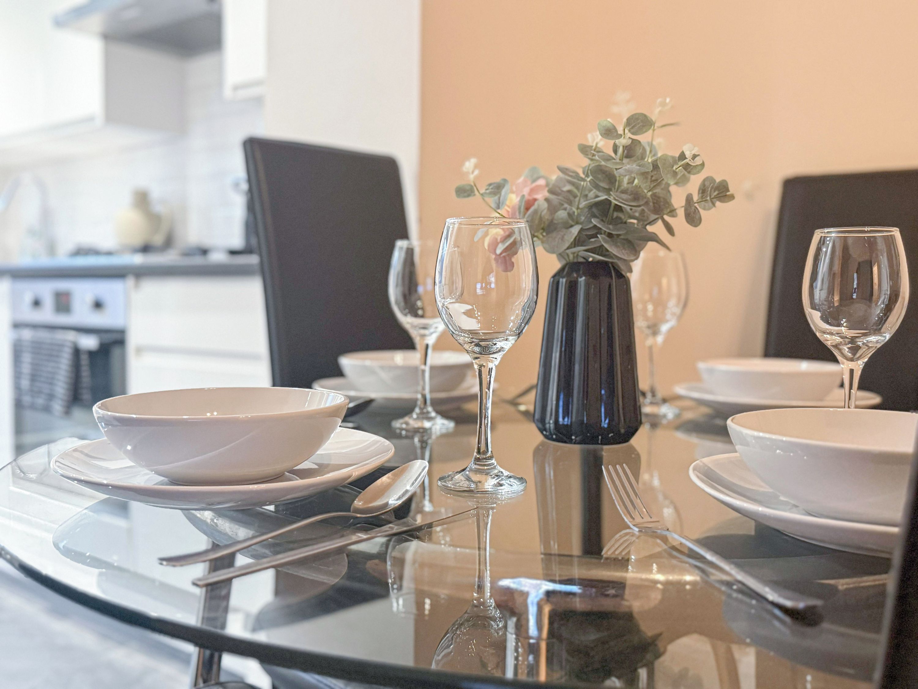Une table à manger dressée avec de l’assiette blanche, des bols, des couverts et des verres à vin, et un vase rempli de fleurs placé au centre.