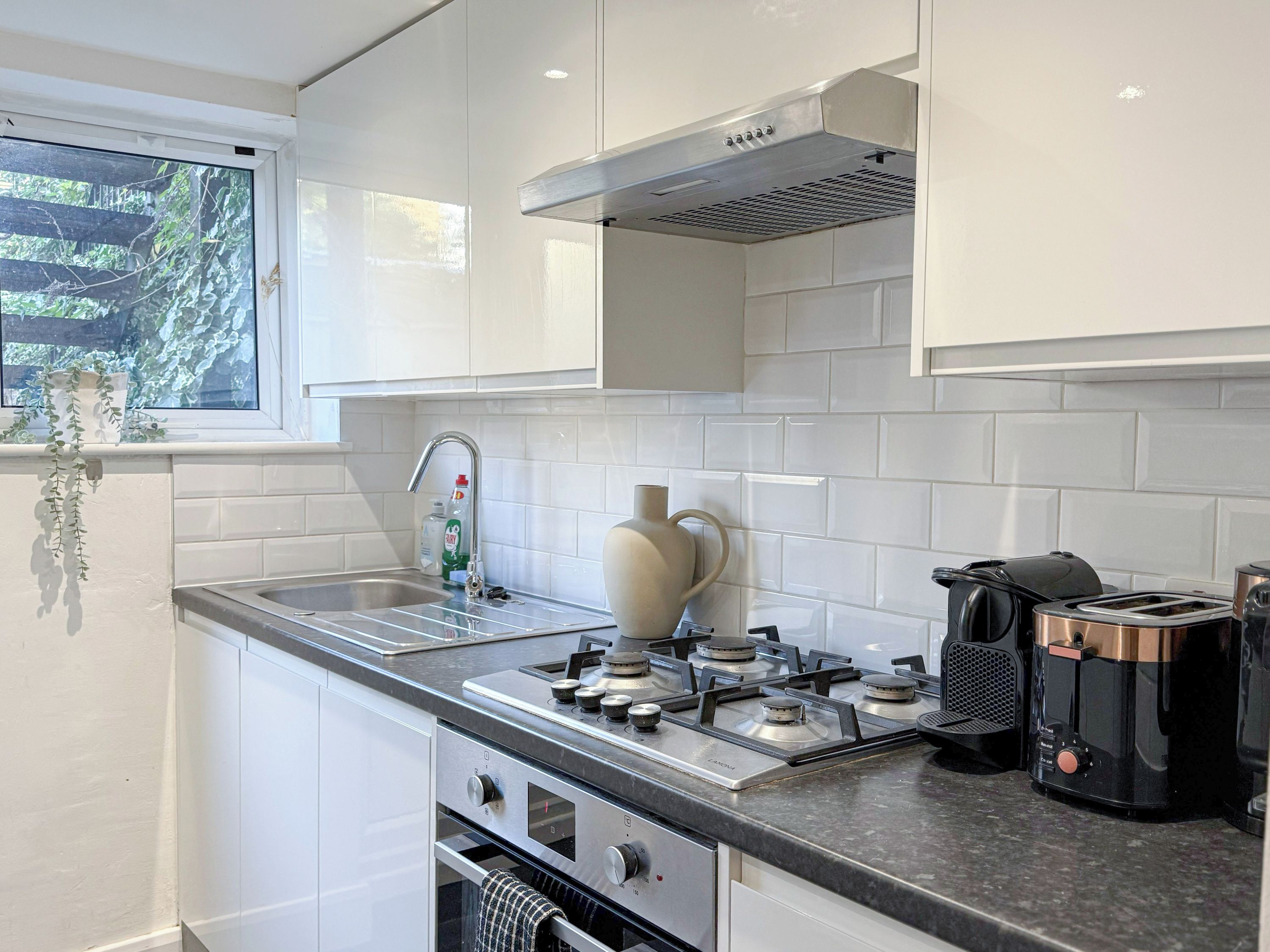 Een moderne keuken met witte kasten, een roestvrijstalen fornuis, een inbouwoven, een raam met uitzicht op groen en een koffiezetapparaat.