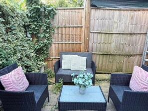 A patio area with wicker furniture and a small table, surrounded by a wooden fence and greenery.
