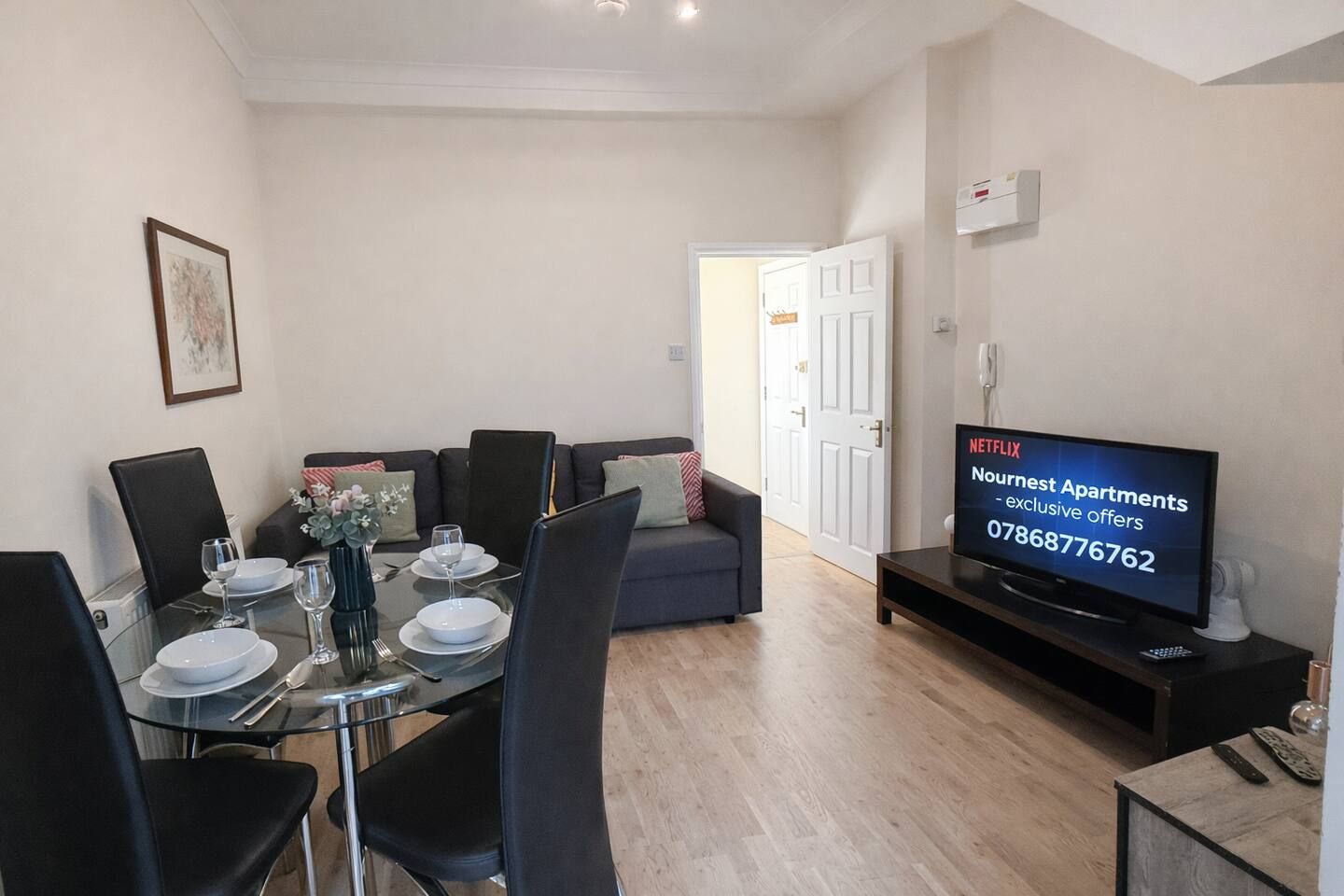 A modern living room with a dining table set for four, a sofa, a television, and a door leading to another room.