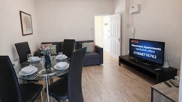 A modern living room with a dining table set for four, a sofa, a television, and a door leading to another room.