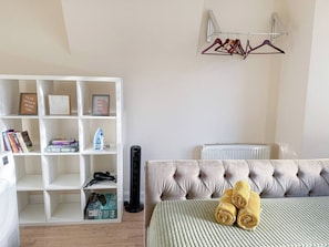 Un dormitorio con cabecera capitonada, una estantería blanca con libros y un altavoz negro, y un radiador.