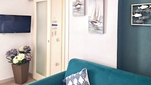 A room with a teal sofa, a wall-mounted TV, and three framed artworks depicting sailboats.