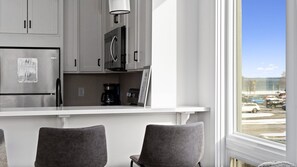 A modern kitchen with grey cabinets, a stainless steel refrigerator, a coffee maker, and a view of a parking lot through a window. - Summer Break! Downtown | Lake Views! NEW 4BR (Traverse City)