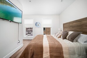 A bedroom with a large bed, a mounted TV, a window with blinds, and a view of a cityscape. - Summer Break! Downtown | Lake Views! NEW 4BR (Traverse City)