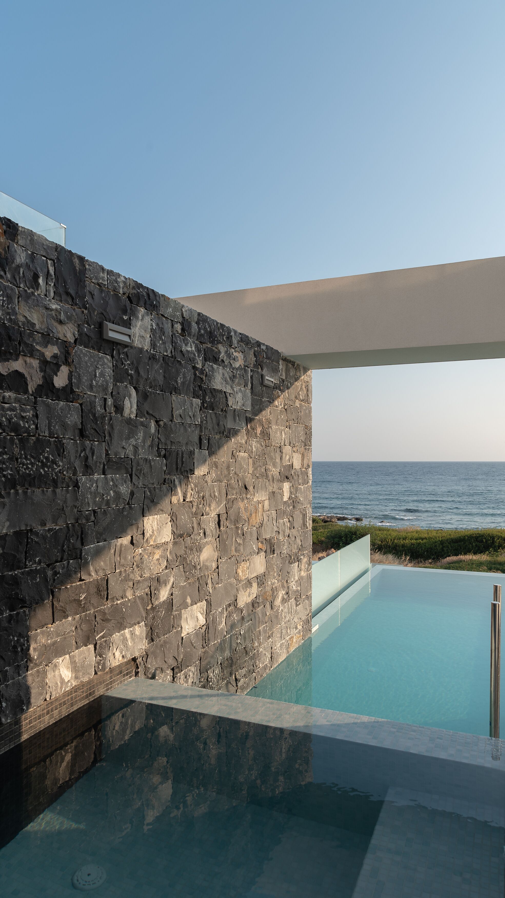 A modern pool area with a stone wall and a view of the sea.