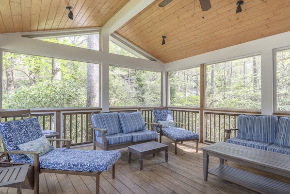 A covered porch with wooden flooring, multiple seating areas with patterned cushions, and large windows offering a view of a forested area.