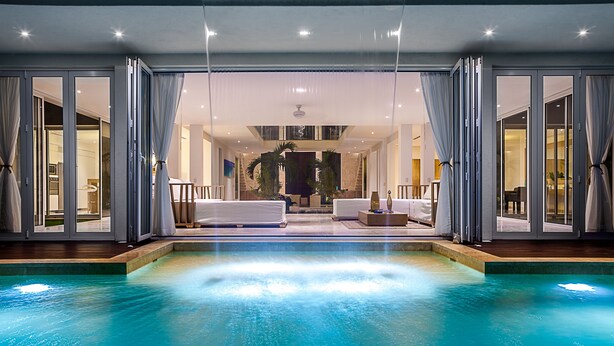 En moderne swimmingpool indendørs med klart vand, omgivet af glasdøre og gardiner, der fører til et godt oplyst opholdsområde med sofaer og pejs.