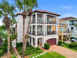 A multi-story residential building with a two-story balcony, a garage, and a well-maintained garden with palm trees.