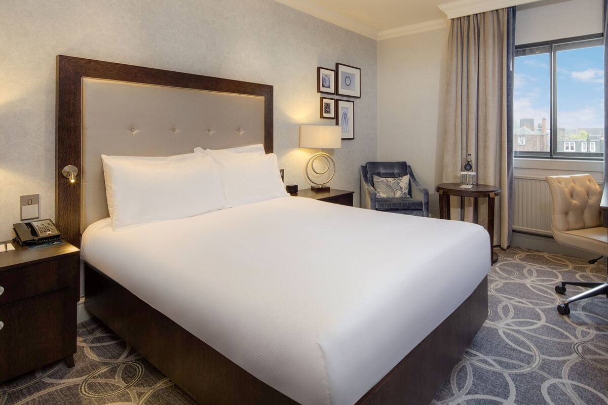 Ein Hotelzimmer mit einem großen Bett, einem Schreibtisch, einem Stuhl, einem Nachttisch mit Telefon und gerahmten Bildern an der Wand.