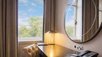 A hotel room with a desk, a lamp, a telephone, and a view of trees through the window.
